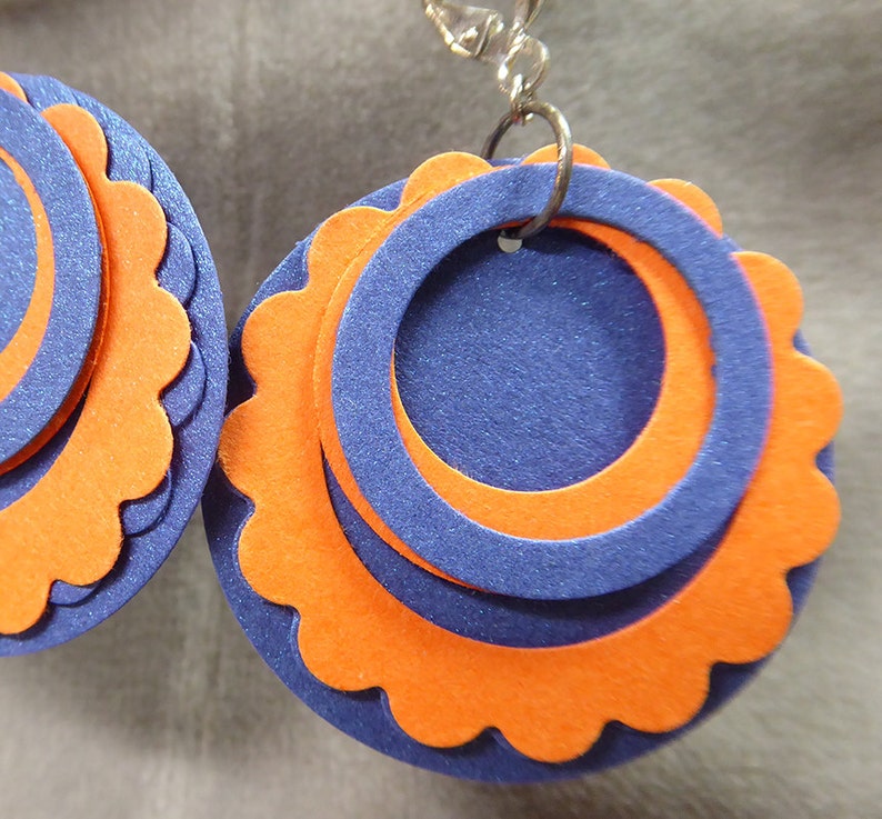 May include: A pair of earrings with a layered design. The earrings are made of orange and blue felt and have a scalloped edge. The earrings are attached to silver hooks.