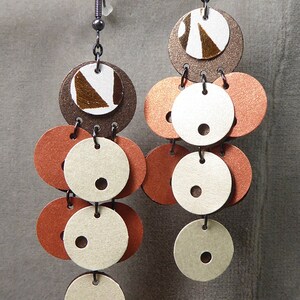May include: A pair of dangle earrings with multiple layers of brown, white, and orange circles. The circles are made of leather and have a textured finish.