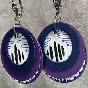 May include: A pair of dangle earrings with a layered design. The earrings are made of purple and navy blue felt with a white and black patterned circle in the center. The earrings have silver hooks.