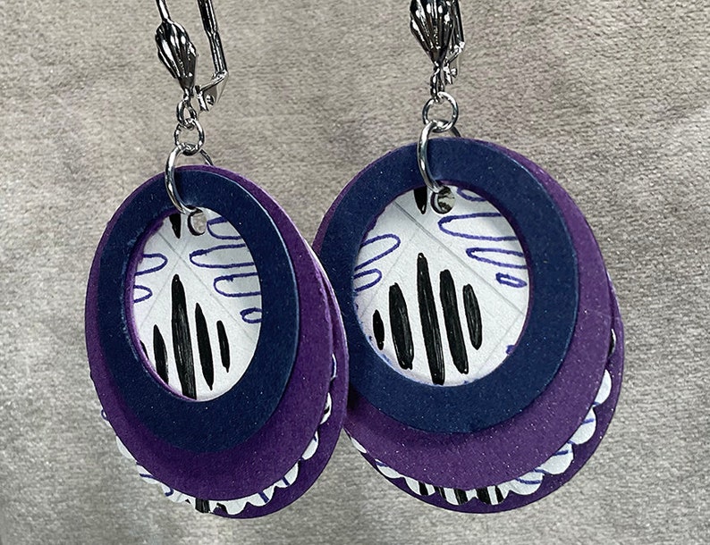 May include: A pair of dangle earrings with a layered design. The earrings are made of purple and navy blue felt with a white and black patterned circle in the center. The earrings have silver hooks.