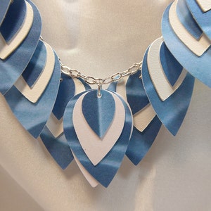 May include: A blue and white layered paper necklace with a silver chain. The necklace is made of multiple layers of paper, each with a different shade of blue. The chain is attached to the top layer of paper.