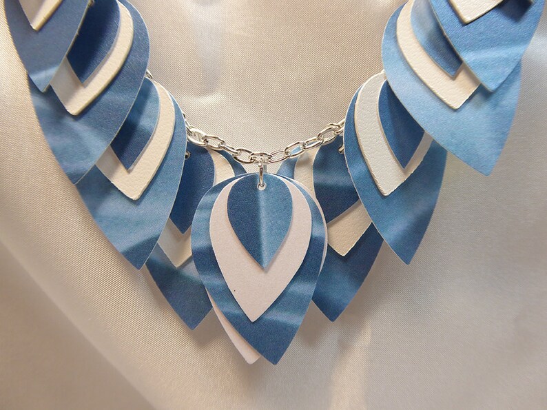 May include: A blue and white layered paper necklace with a silver chain. The necklace is made of multiple layers of paper, each with a different shade of blue. The chain is attached to the top layer of paper.