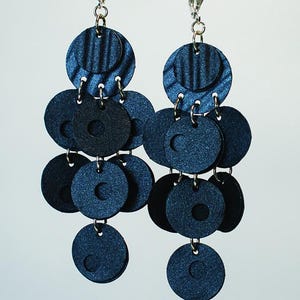 Op de afbeelding: Een paar hangende oorbellen met meerdere donkerblauwe ronde schijven van verschillende groottes, verbonden door zilverkleurige metalen ringetjes. De bovenste schijven hebben een geribbelde textuur, terwijl de onderste schijven glad en glinsterend zijn.