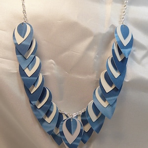 May include: A blue and white paper scale necklace with a silver chain. The necklace is made of overlapping teardrop-shaped pieces of paper.