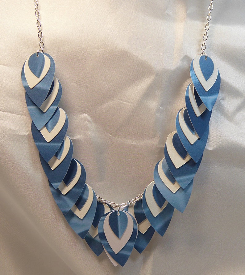 May include: A blue and white paper scale necklace with a silver chain. The necklace is made of overlapping teardrop-shaped pieces of paper.
