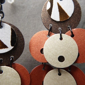 May include: A pair of dangle earrings with multiple layers of brown, white, and orange circles. The top circle has a brown, white, and gold geometric pattern.