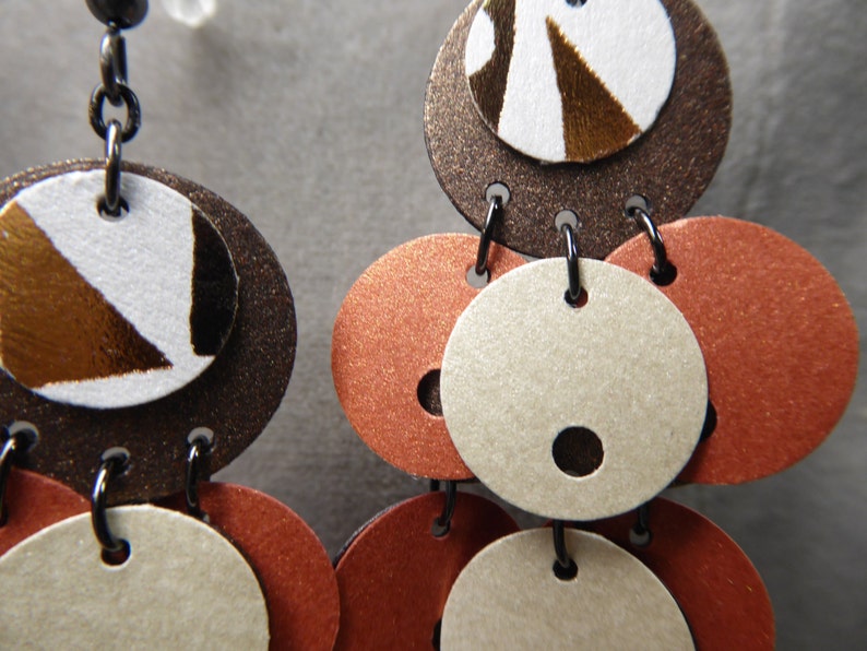 May include: A pair of dangle earrings with multiple layers of brown, white, and orange circles. The top circle has a brown, white, and gold geometric pattern.