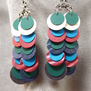 May include: A pair of dangle earrings with multiple layers of round discs in shades of green, blue, red, and white. The discs are layered in a graduated size, with the largest disc at the bottom. The earrings are attached to silver-toned hooks.