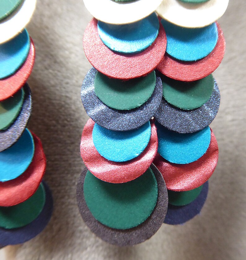 May include: A pair of earrings with multiple layers of colorful circles. The circles are in shades of teal, green, red, and black. The earrings are made of a shiny material.