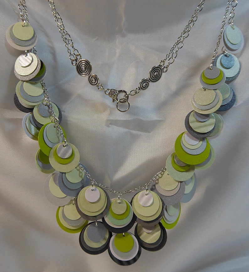 May include: A silver chain necklace with a silver clasp. The necklace is adorned with layered circles of green, gray, and white paper.