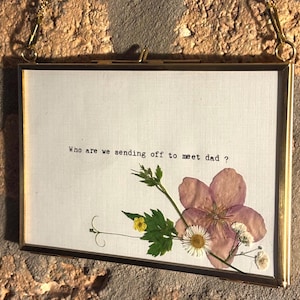 May include: A rectangular glass frame with a thin gold metal border and a hanging chain. Inside, a white fabric background features the black printed text: Who are we sending off to meet dad? Pressed dried flowers include a large pink blossom.