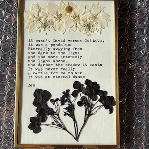Könnte beinhalten: Ein gerahmtes Kunstwerk mit gepressten Blumen und einem Gedicht. Der goldfarbene Rahmen enthält einen weißen Hintergrund mit getrockneten weißen Blumen oben und dunklen Blumen unten. Das Gedicht ist in schwarzem Text gedruckt.