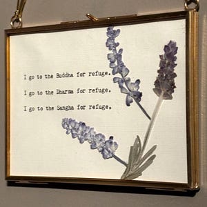 May include: A rectangular framed art piece with a gold-colored metal frame. The artwork features dried lavender sprigs and the text "I go to the Buddha for refuge. I go to the Dharma for refuge. I go to the Sangha for refuge." The background is off-white.