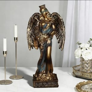 Könnte beinhalten: Eine bronzefarbene Statue einer geflügelten Figur, möglicherweise eines Engels oder einer Göttin, die ein Füllhorn hält. Die Figur ist mit verbundenen Augen und trägt eine Krone. Zwei goldene Kerzenhalter mit weißen Kerzen befinden sich links.