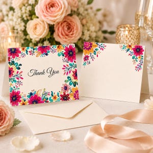 May include: Two folded cream-colored cards with a vibrant floral border featuring pink, yellow, and teal watercolor flowers. One card displays the text Thank You in black script. An empty cream envelope and a satin ribbon are positioned nearby.