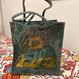 Op de afbeelding: Een vintage groene Marshall Field's boodschappentas met een glanzende, gemarmerde afwerking. De tas heeft een gouden logo en belettering, met bijpassende groene handvatten. De tas staat op een oppervlak met bloemenpatroon.