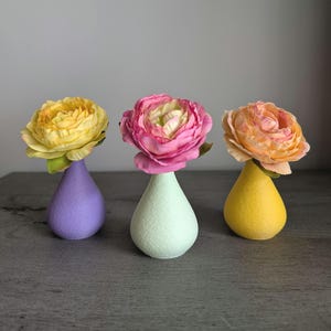 May include: Three artificial flowers in small, teardrop-shaped vases. The left vase is purple with a yellow flower, the center vase is mint green with a pink flower, and the right vase is yellow with a peach-colored flower. Each vase has a textured finish.