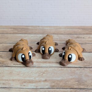May include: Three brown platypus figurines with large white eyes and dark brown beaks and feet are arranged on a wooden surface. The platypuses are lying down, facing forward. The background is a white brick wall.