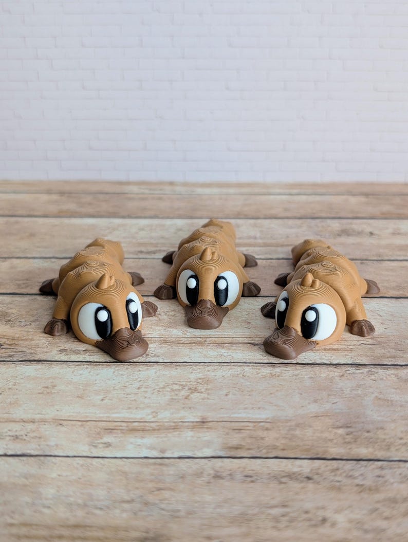 May include: Three brown platypus figurines with large white eyes and dark brown beaks and feet are arranged on a wooden surface. The platypuses are lying down, facing forward. The background is a white brick wall.