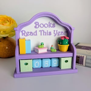 May include: A purple bookshelf-shaped book tracker with the text "Books Read This Year." It features miniature books, a cactus, and two green worm figures. The bottom shelf has cubes with numbers and two green boxes.