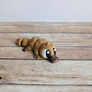 May include: A brown 3D-printed platypus figurine. The figurine has a detailed design with a duck-like bill, large eyes, and a segmented body. The platypus is lying down on a wooden surface.