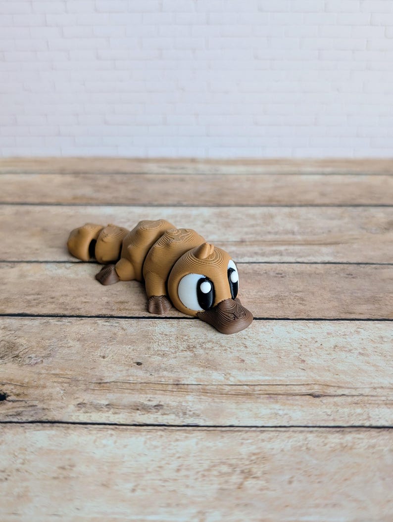 May include: A brown 3D-printed platypus figurine. The figurine has a detailed design with a duck-like bill, large eyes, and a segmented body. The platypus is lying down on a wooden surface.