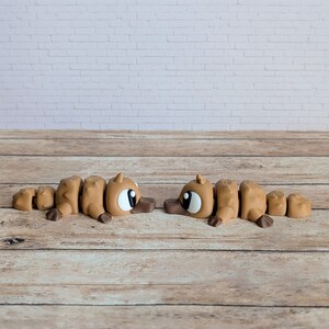May include: Two brown articulated platypus toys with large white eyes and brown beaks are positioned on a wooden surface. The toys are segmented, allowing for movement. The background features a white brick wall.