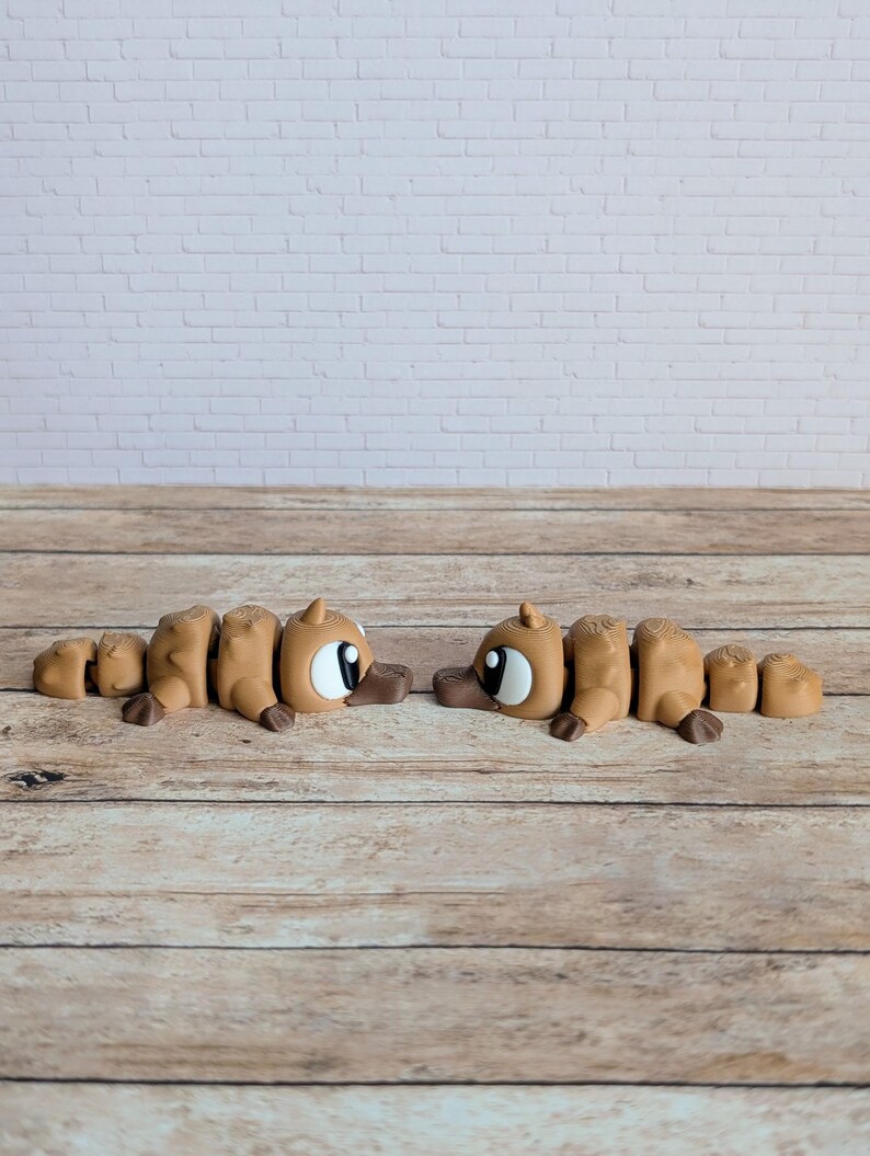 May include: Two brown articulated platypus toys with large white eyes and brown beaks are positioned on a wooden surface. The toys are segmented, allowing for movement. The background features a white brick wall.