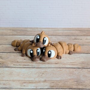 May include: Three brown platypus-shaped figurines with large, expressive eyes and dark brown beaks are arranged on a wooden surface. The figurines are lying down, with their bodies segmented. The background features a white brick wall.