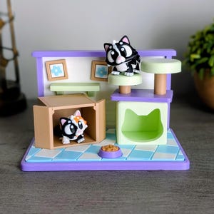 May include: A miniature cat playhouse with two toy cats. The playhouse has a purple roof and base, with a blue and white checkered floor. One cat is inside a cardboard box, and the other is on a green platform. A small bowl of food is also present.