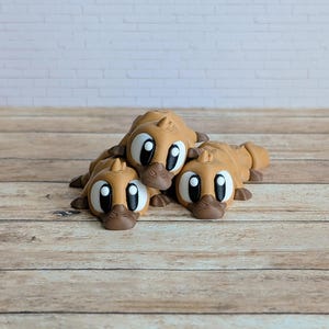 May include: Three brown, cartoon-style platypus figurines with large, expressive eyes and dark brown beaks. The figurines are lying down on a wooden surface, creating a playful and whimsical scene. The background is a light-colored brick wall.