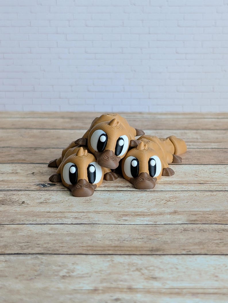 May include: Three brown, cartoon-style platypus figurines with large, expressive eyes and dark brown beaks. The figurines are lying down on a wooden surface, creating a playful and whimsical scene. The background is a light-colored brick wall.