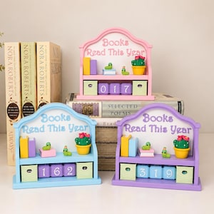 May include: Three wooden bookshelf-shaped trackers with the text "Books Read This Year" printed on the top arch. Each unit features miniature books, a potted cactus, and small green caterpillars, with three numbered blocks to display the total count.