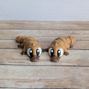 May include: Two brown platypus figurines with large, expressive eyes and a detailed, textured design. The figurines are lying on a wooden surface, showcasing their unique shape and features. The platypuses have a cartoonish appearance.