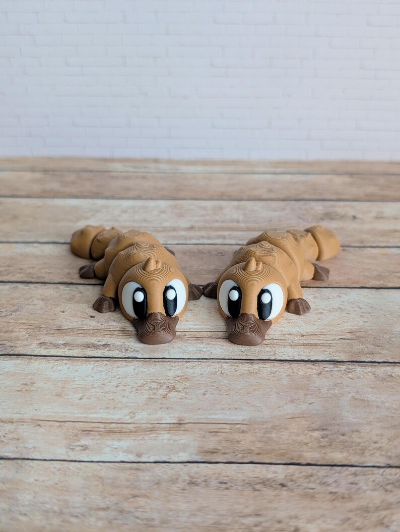 May include: Two brown platypus figurines with large, expressive eyes and a detailed, textured design. The figurines are lying on a wooden surface, showcasing their unique shape and features. The platypuses have a cartoonish appearance.