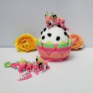 May include: A white egg-shaped container with black spots, encased in a pink and green petal-like base. A small pink and white dragon figure with green horns rests on top, while a matching articulated dragon figure lies on the surface nearby.