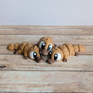 May include: Three brown platypus figurines with large, expressive eyes and dark brown beaks are arranged on a wooden surface. The figurines are lying down, showcasing their detailed design and playful appearance. The background features a light-colored brick wall.