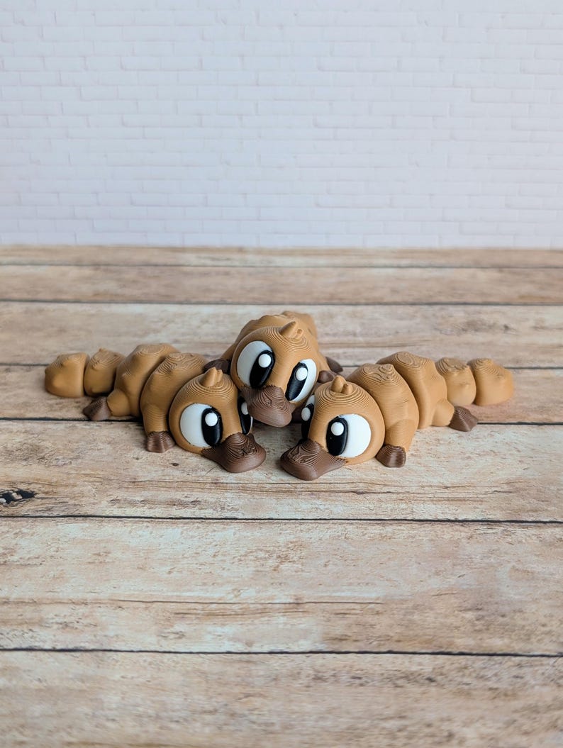 May include: Three brown platypus figurines with large, expressive eyes and dark brown beaks are arranged on a wooden surface. The figurines are lying down, showcasing their detailed design and playful appearance. The background features a light-colored brick wall.