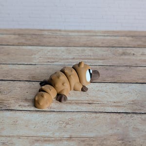 May include: A segmented, light brown platypus figurine with dark brown feet and a beak. The toy has a white eye with a black pupil. The figurine is lying on a wooden surface with a white brick wall in the background.
