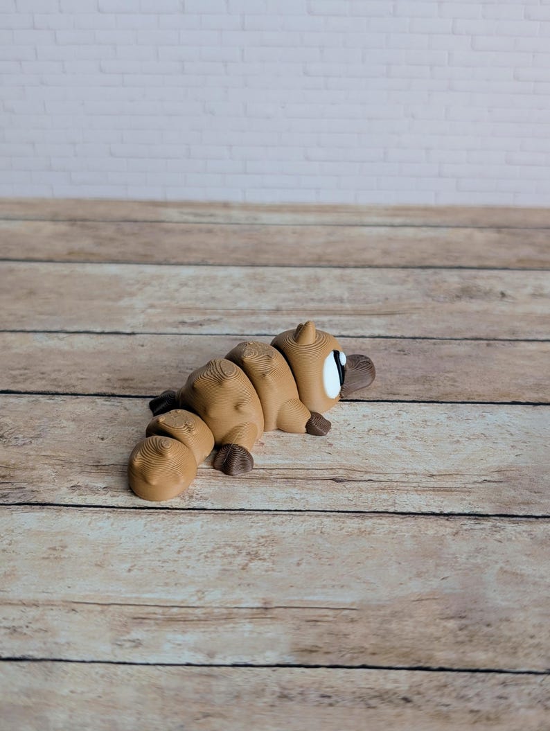 May include: A segmented, light brown platypus figurine with dark brown feet and a beak. The toy has a white eye with a black pupil. The figurine is lying on a wooden surface with a white brick wall in the background.
