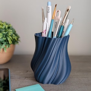 May include: A blue, 3D printed vase with a swirling design holds a variety of art supplies, including paintbrushes, pens, and markers. The vase is sitting on a wooden surface with a light blue sticky note and a white pen.