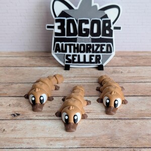 May include: Three brown platypus figurines with large white eyes and dark brown beaks are displayed on a wooden surface. A sign with the text "3DGOB AUTHORIZED SELLER" is in the background.