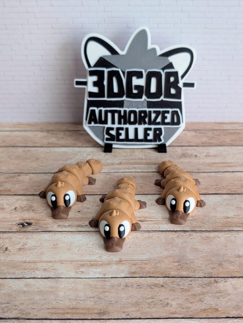 May include: Three brown platypus figurines with large white eyes and dark brown beaks are displayed on a wooden surface. A sign with the text "3DGOB AUTHORIZED SELLER" is in the background.