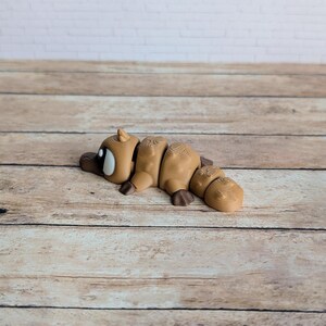 May include: A segmented, tan-colored platypus figurine with dark brown feet and a dark brown bill. The figurine has a white and black eye and is lying on a wooden surface. The background is a white brick wall.