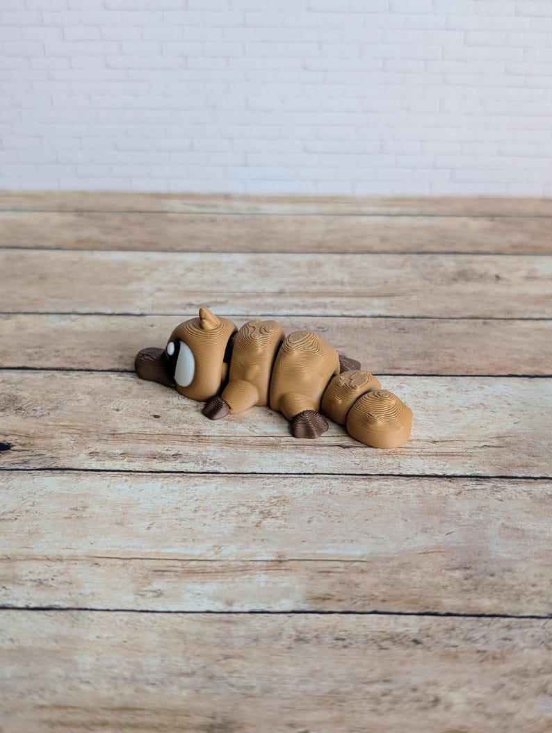 May include: A segmented, tan-colored platypus figurine with dark brown feet and a dark brown bill. The figurine has a white and black eye and is lying on a wooden surface. The background is a white brick wall.