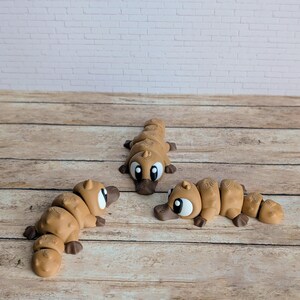 May include: Three tan-colored, segmented platypus toys with large eyes and dark brown beaks are arranged on a wooden surface. The toys are in various poses, with one lying down and two upright. A white brick wall is in the background.