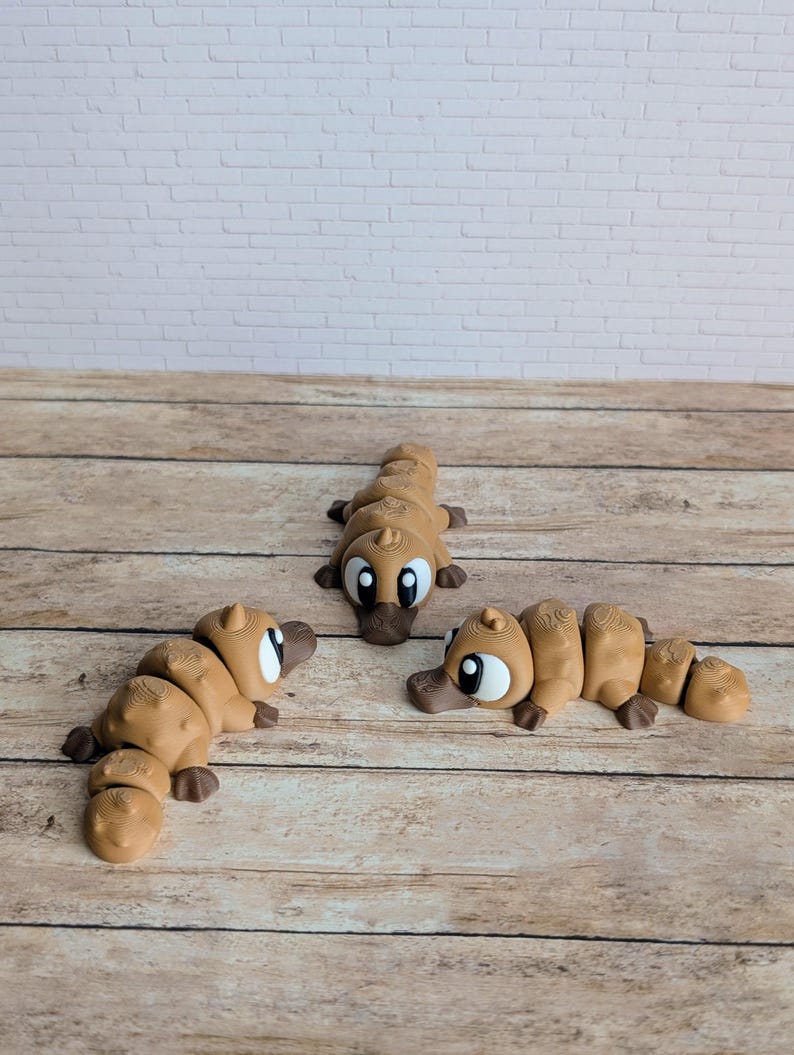 May include: Three tan-colored, segmented platypus toys with large eyes and dark brown beaks are arranged on a wooden surface. The toys are in various poses, with one lying down and two upright. A white brick wall is in the background.