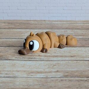 May include: A brown 3D-printed platypus toy with large white eyes and a dark brown bill. The toy is segmented, with a rounded body and a small tail. It is lying on a wooden surface.