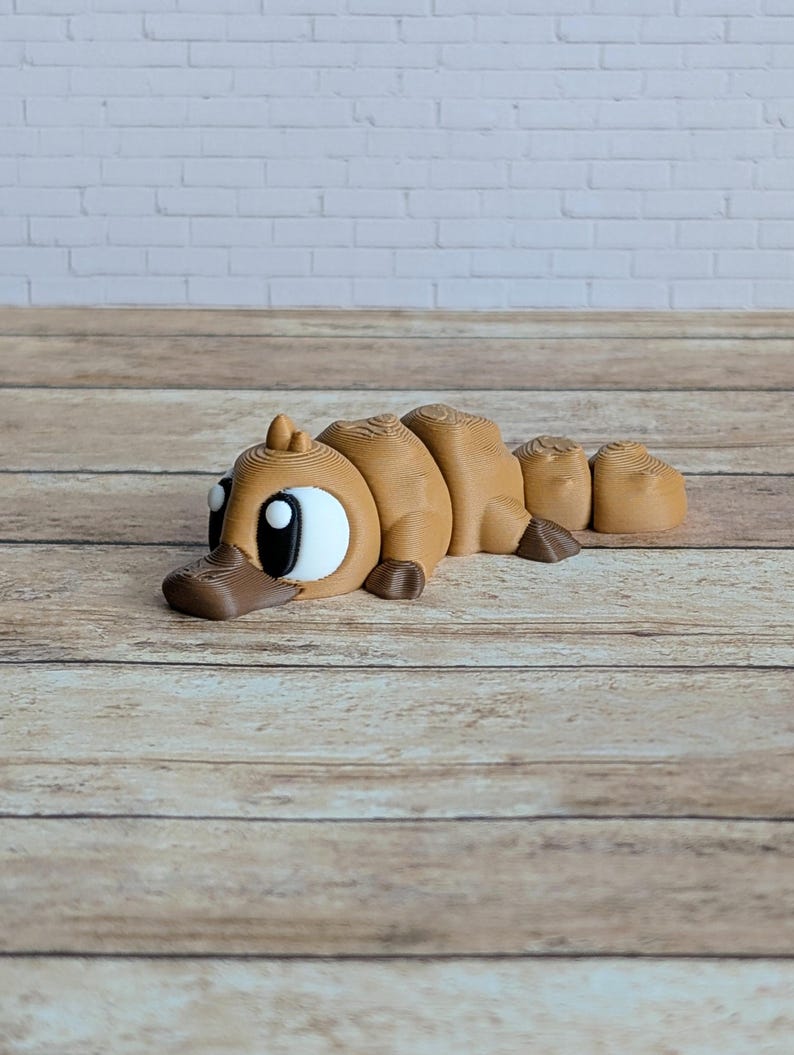 May include: A brown 3D-printed platypus toy with large white eyes and a dark brown bill. The toy is segmented, with a rounded body and a small tail. It is lying on a wooden surface.