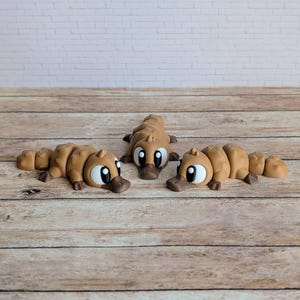 May include: Three brown platypus figurines with large eyes and duck-like bills are arranged on a wooden surface. The figurines are lying down, with their bodies segmented. The background features a white brick wall.